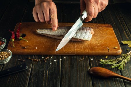 Un cocinero usa un cuchillo para cortar pescado fresco de merluza en una tabla de cortar de madera. Un concepto bajo en clave para preparar un plato nacional de pescado en casa utilizando una receta única.