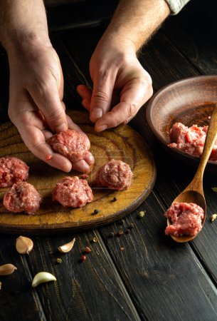 The chef shapes minced meat into homemade meatballs for lunch. Low key concept of cooking food on kitchen table with meat, vegetables and spices. Advertising space.