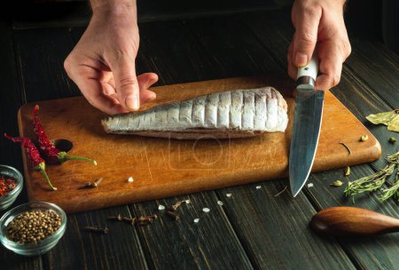 The cook is preparing sea fish on the kitchen table. Knife in the hand of a chef for slicing fish. Low key concept of fish dish for lunch.