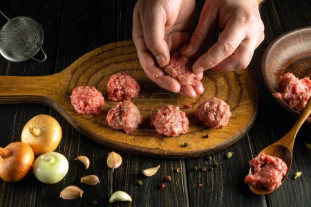 The cook uses his hands to form minced meat to prepare meatballs for breakfast. Low key concept of cooking food on kitchen table with vegetables and spices.