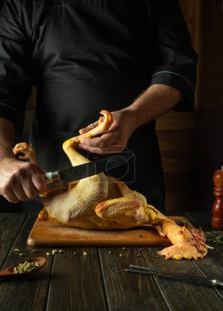 Chef cuts raw rooster with a knife in the saloon kitchen. Concept of preparing a chicken dish for lunch on a dark background.