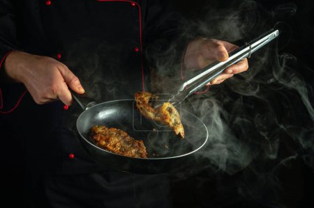 Ein professioneller Koch entfernt mit einer Zange heißen gebratenen Fisch aus der Pfanne. Low-Key-Konzept der Zubereitung eines köstlichen Fischgerichts in einer Restaurant- oder Kneipenküche.