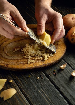 Auf dem Küchentisch bereitet der Koch Kartoffelpuffer zu. Großaufnahme eines Mannes, der rohe Kartoffeln mit einer Reibe reibt.