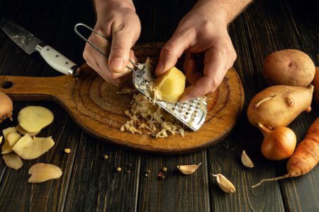 Die Hände eines Kochs mit einer Reibe reiben rohe Kartoffeln auf dem Küchentisch. Konzept der Zubereitung von leckerem Gemüse-Frühstück.