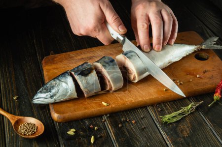 The cook cuts raw fish with a knife before baking it on the grill. Spices on the kitchen table for preparing a delicious breakfast with mackerel.