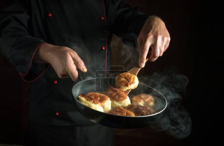 A professional chef fries donuts in a restaurant kitchen. Kitchen spatula in the hand of the cook and a hot frying pan. Low key concept of making delicious pancakes.