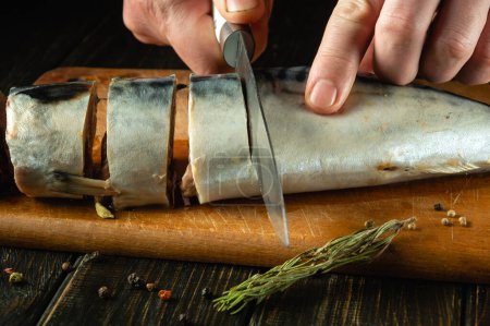 Primer plano de las manos de un chef con un cuchillo rebanando caballa antes de cocinar. Concepto Low Key para menú de pescado con especias y romero.