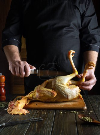 Foto de Cortar pollo crudo con un cuchillo en la mano del chef en la mesa de la cocina para asar para el almuerzo. - Imagen libre de derechos