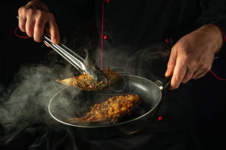 Der Koch brät in der Küche Fisch. Das Konzept, ein Fischgericht zum Mittagessen zuzubereiten. Die Köchin holt mit einer Zange heiße gebratene Karauschen aus der Pfanne.