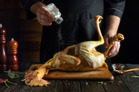 A cook prepares a raw rooster on the kitchen table in a restaurant. Before roasting, the chef adds salt to the chicken.