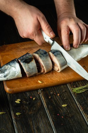 Slicing raw fish for salting with spices with a knife in the hands of a cook. The concept of preparing herring with fish on the kitchen table.