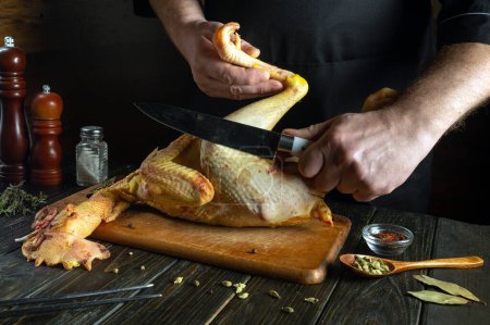 Foto de Cocinar gallo crudo en la mesa de la cocina. Un cuchillo en la mano del chef para cortar pollo para preparar la cena. - Imagen libre de derechos