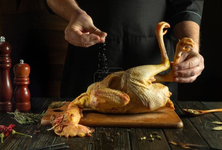 Foto de Antes de asar, el gallo debe ser salado. El cocinero agrega sal al pollo crudo en la mesa de la cocina. - Imagen libre de derechos