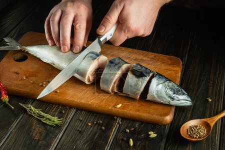 The chef cuts raw fish with a knife before salting it. Spices on the kitchen table for preparing a delicious dinner with mackerel.
