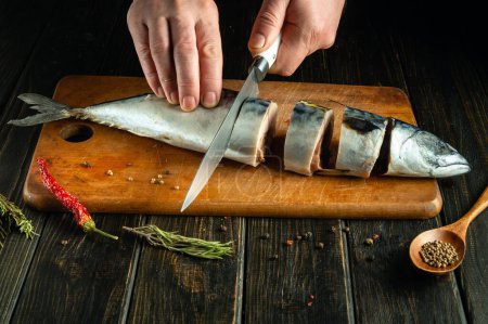 Slicing raw mackerel fish on a kitchen board with a knife in the chef's hand before salting with spices.