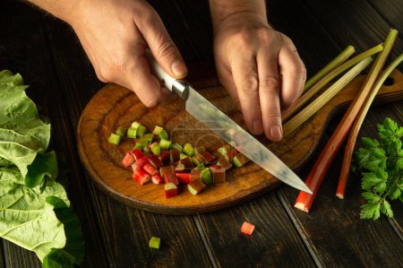 Preparing a vegetarian dish from rheum plant. Chef hands use a knife to slicing rhubarb stalks on a kitchen board before preparing dinner.