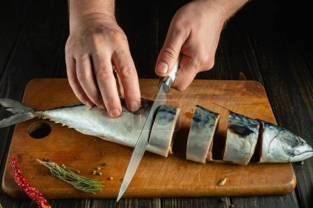 The chef hands cut the mackerel into pieces with a knife before frying it for dinner. Working environment on a kitchen table in a restaurant.