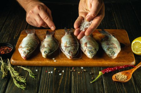 Primer plano de las manos de un chef añadiendo sal gruesa al pescado crudo en una tabla de cortar. El concepto de cocinar carpa cruciana salada con especias y cilantro en la mesa de la cocina.