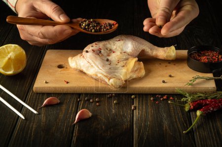 Close-up of a chef hands with a spoon adding pepper to a chicken leg. Broiler leg on cutting board with rosemary and spices before grilling.