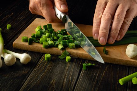 Der Koch schneidet grünen jungen Knoblauch auf einem hölzernen Schneidebrett mit einem Messer für die Zubereitung vegetarischer Speisen. Werbefläche.