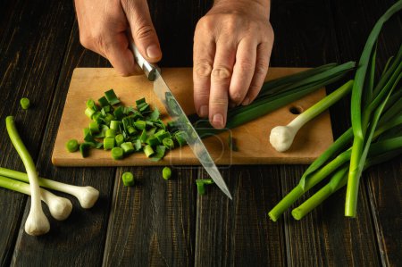 Chef Hände mit einem Messer, um grünen Knoblauch auf einem Schneidebrett für die Zubereitung eines vegetarischen Gerichts oder Salats schneiden. Platz für Werbung.