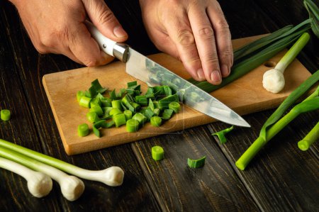 Kochen eines schmackhaften vegetarischen Gerichts mit Knoblauch. Die Hände des Küchenchefs schneiden jungen Knoblauch mit einem Messer auf ein hölzernes Küchenbrett.