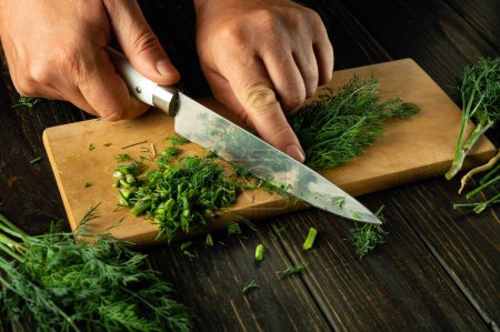 A chef uses a knife to chop fresh green dill on a wooden board before adding it to a vegetarian dish. Space for advertising on a black table.