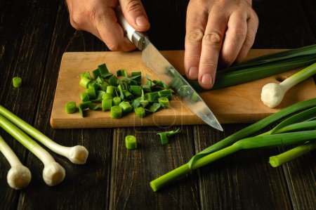 A cook cuts green garlic on a cutting board with a knife to prepare a vegetarian dish. Peasant food.