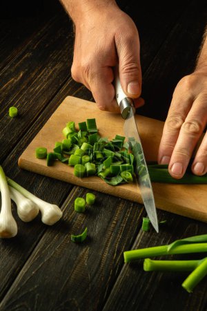 A cook cuts green garlic on a cutting board with a knife to prepare a vegetarian dish. Place for advertising.