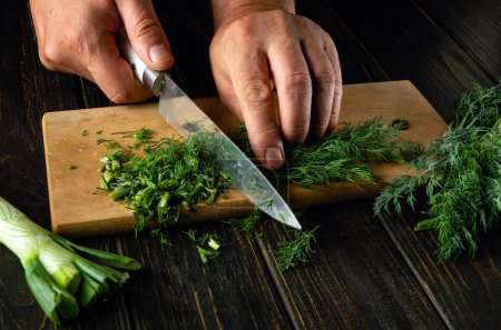 Chef hands use a knife to cut fresh dill on a cutting board for preparing a vegetarian dish. Peasant products on the kitchen table.