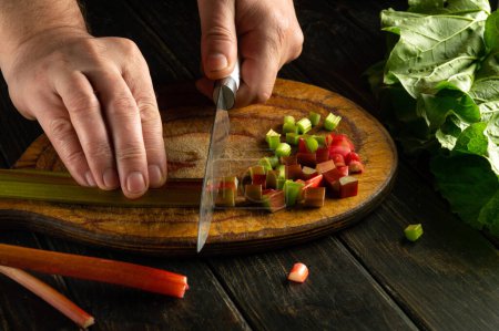 A cook uses a knife to cut rhubarb stalks on a wooden board to prepare a vegetarian meal.