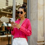 Adorable stylish young caucasian woman spends time at weekend in the city. Brunette wears sunglasses, pink shirt and shorts. She scrolling smartphone. Lifestyle concept