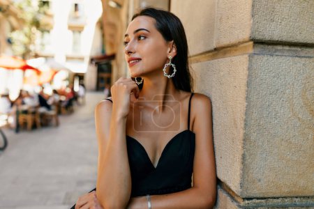 Photo for Elegant adorable stylish woman with tanned skin and loose dark hair wearing black dress and festive earrings is posing near the wall on the street and looking aside - Royalty Free Image