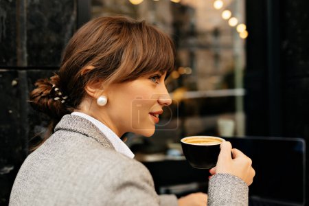 Close up portrait from back dark-haired woman drinking coffee on city background with lights. Charming woman in grey jacket smiling and chilling outside.