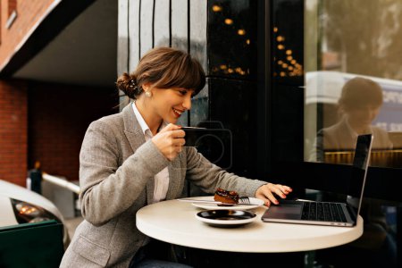 Photo for Happy smiling woman in gray jacket and white blouse working on laptop with coffee. Remote work, good mood. - Royalty Free Image