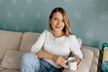 Pretty charismatic girl with short blond hair wearing white shirt and jeans looking at camera with lovely smile, holding coffee and posing at camera.
