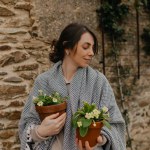 Close up profile portrait of cute smiling lady with dark hair throws plaid on shoulders is holding flowers in hand, looking aside and smiling with closed eyes on background of stone house 