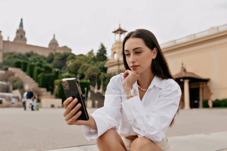 Elegant cute woman with dark loose hair wearing white shirt, using smartphone and resting outdoor. Brunette wears casual clothes. Weekend concept, technology. 