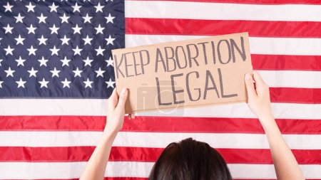 Foto de Woman is holding an empty cardboard with Space for Text sign US flag on background. Protest against anti abortion law. Women's strike. Womens rights freedom. Against racism racial discrimination. - Imagen libre de derechos