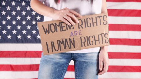 Téléchargez les photos : Femme méconnaissable tient une pancarte avec signe Les droits des femmes sont des droits de l'homme dans la main drapeau américain arrière-plan. Une fille tient du carton avec inscription. Manifestation des droits des femmes - en image libre de droit