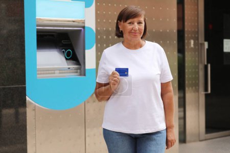 Photo for Beautiful middle aged woman is showing credit card near bank cash machine outdoors. Mature female withdrawing money with debit card in ATM - Concept of business, banking account and lifestyle people. - Royalty Free Image