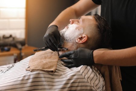 Photo for Professional barber in black gloves is shaving client with a vintage straight stainless steel razor in barbershop. Shaving cream, foam. Hairdresser, beauty and fashion salon. - Royalty Free Image