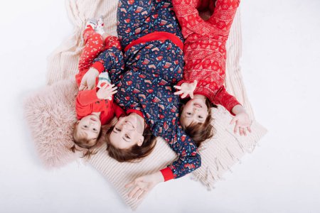 Téléchargez les photos : Vue du dessus de maman souriante avec deux petites filles en pyjama d'hiver dans les impressions de Noël sont couchés sur le plaid chaud. Une famille heureuse s'étreint et s'amuse ensemble. Fête des mères, fête des bébés. Nouvel An vacances. - en image libre de droit