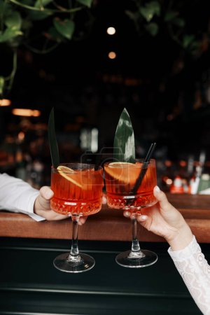 hands of woman and man are clinking, cheers with glasses of Spritz cocktail. Couple celebrating wedding, anniversary with Aperol spritz cocktails, with orange and greens Refreshing alcoholic drink.