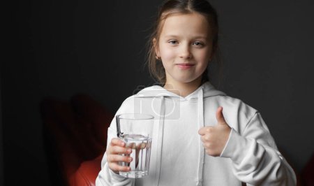 Little girl drinks fresh transparent pure filtered mineral water from glass and shows thumbs up at home. Thirsty dehydrated teenager enjoys fresh drink. Water balance and children health care
