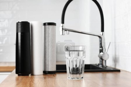 A glass of clean fresh water and set of filter cartridges on wooden table in a kitchen interior. Installation of reverse osmosis water purification system. Concept Household filtration system