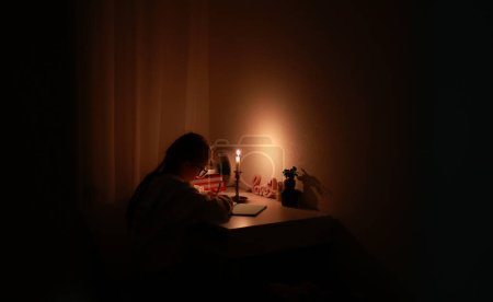 Schoolgirl uses candlelight to writes and reads in darkness without electric lights at home. Teen with poor eyesight wears glasses to studies and to do school homework during blackout.Vision problems.