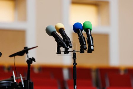 Vier farbige Mikrofone stehen auf dem Podium, im Hintergrund verschwimmen viele rote Stühle. Kommunikationskonzept, Veranstaltung, Seminar, Medien, Pressekonferenz, Performance, Politik, Unternehmen