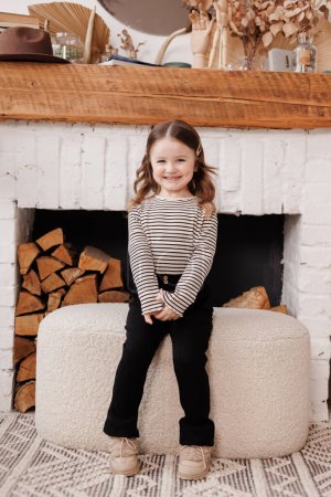 Photo for Charming little girl sits on soft pouffe chair relaxes at living room in Scandinavian style. Child in striped longsleeve looks at camera with happiness and mysterious smile. Domestic comfort concept. - Royalty Free Image