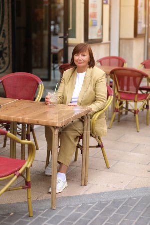 Téléchargez les photos : Attrayant élégant femme mûre 50-60 s avec les cheveux courts en veste jaune est de boire du lait de café boisson avec de la paille et de détente au café dans la rue de la ville européenne. Concept de vacances d'été. - en image libre de droit